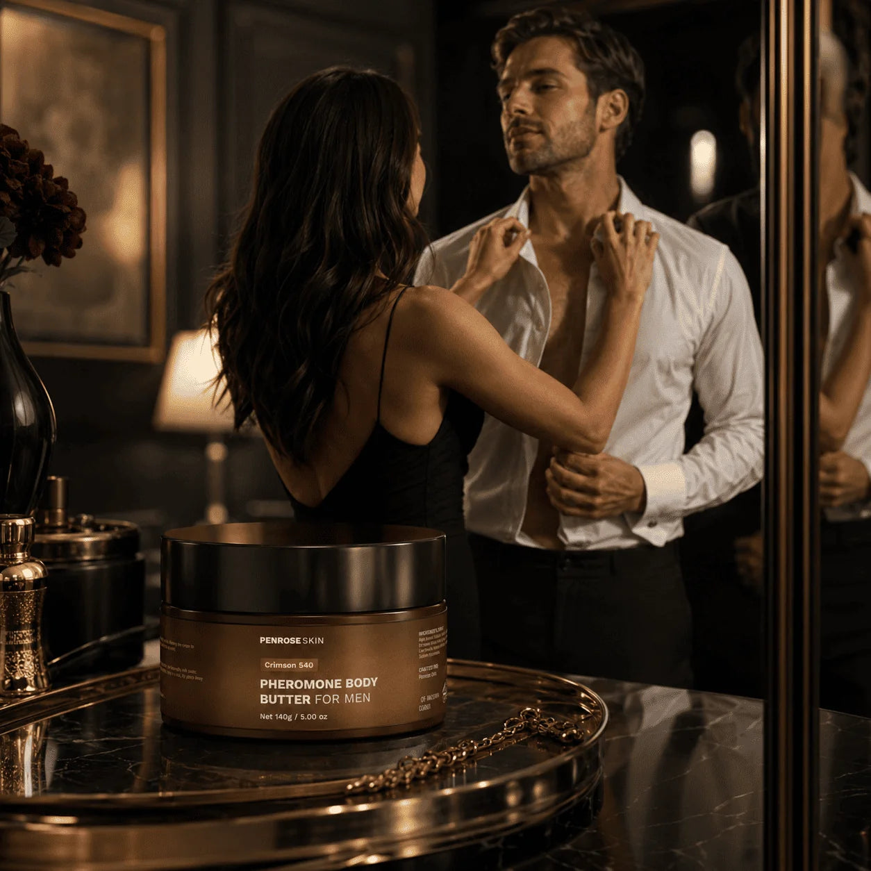 Penrose Skin Crimson 540 pheromone body butter on a gold tray as a couple gets ready in a dark, warmly lit dressing room